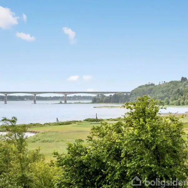 Bro strækker sig over en bred flod, omgivet af frodige grønne træer og buske. Blå himmel med få hvide skyer danner en rolig baggrund.