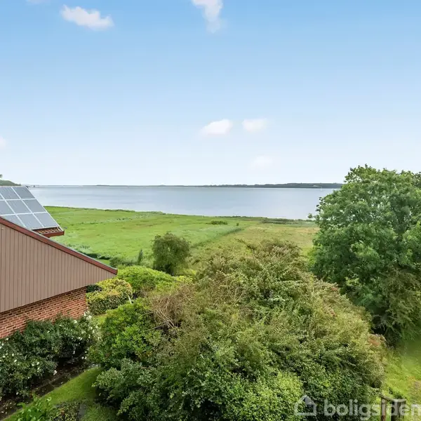 Rødt murstenshus med solpaneler på taget står tæt ved en grøn have. I baggrunden ses en stor sø under en klar, blå himmel, omkranset af frodige træer og græsarealer.