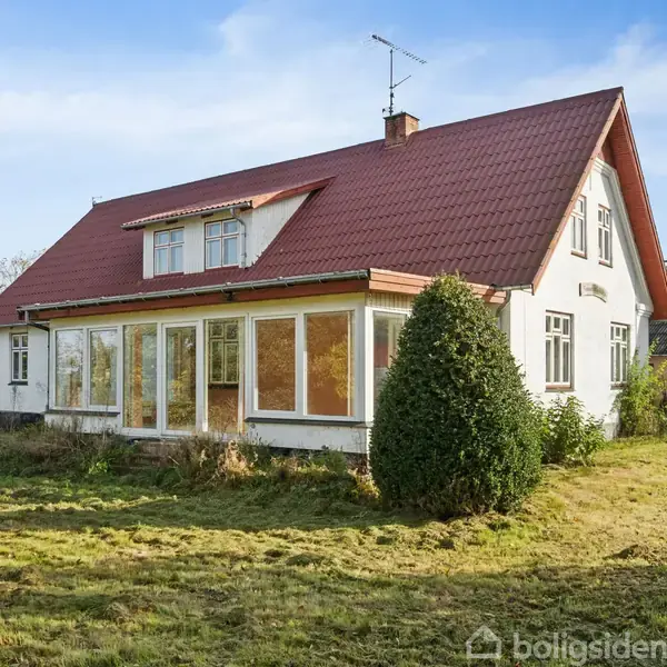 Et hvidt hus med rødt tag står på en åben græsplæne. Huset har store vinduer og omgivet af buske og træer under en klar blå himmel.