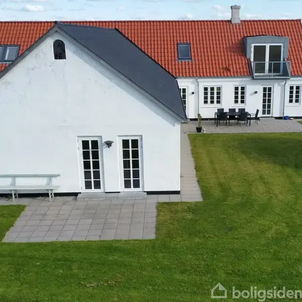 Hvidt hus med rødt tegltag står på en velplejet græsplæne. Huset har udendørs terrasse med bord og stole; omgivet af åbne marker og få andre bygninger i baggrunden.