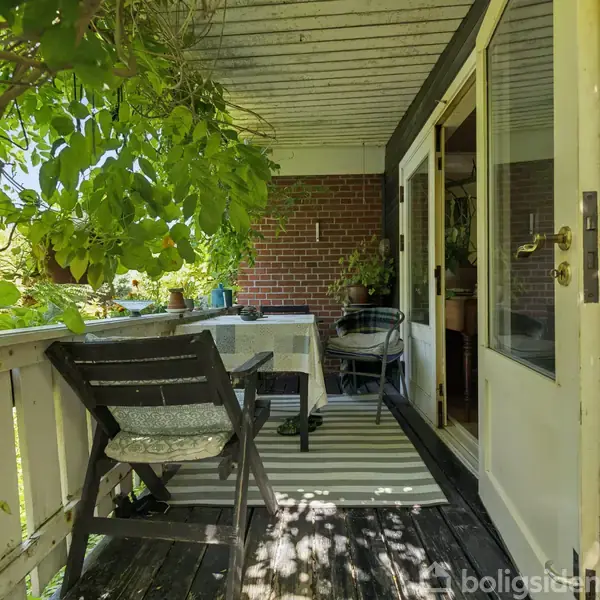 Terrasse med træbord og stole står stille. Omgivet af grønne planter, hvidt rækværk og en åben havedør, skaber den en hyggelig atmosfære.