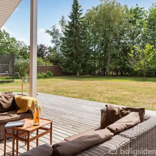 Terrassemøbler står stille på en træterrasse, hvor en sofa og et bord med en flaske og glas skaber en behagelig opholdsplads. Områdets græsplæne og skovbevoksning skaber en naturlig ramme.