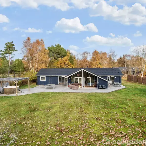 Et moderne sort-hus med en stor terrasse ligger i en åben grøn have omgivet af træer. Terrassen har møbler, og til venstre ses en pergola. Himmel med lette skyer.