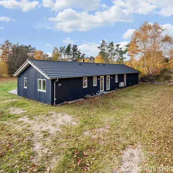 Et mørkeblåt hus står stille på en stor, græsbevokset grund omgivet af efterårstræer med gyldne blade. Nummer 26 er skrevet på husets væg ved siden af en dør.