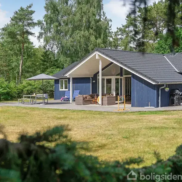 Et blåmalet hus med terrasse står i en grøn have omkranset af træer. Havemøbler og en parasol ses på terrassen. Sollys gennemstrømmer området.