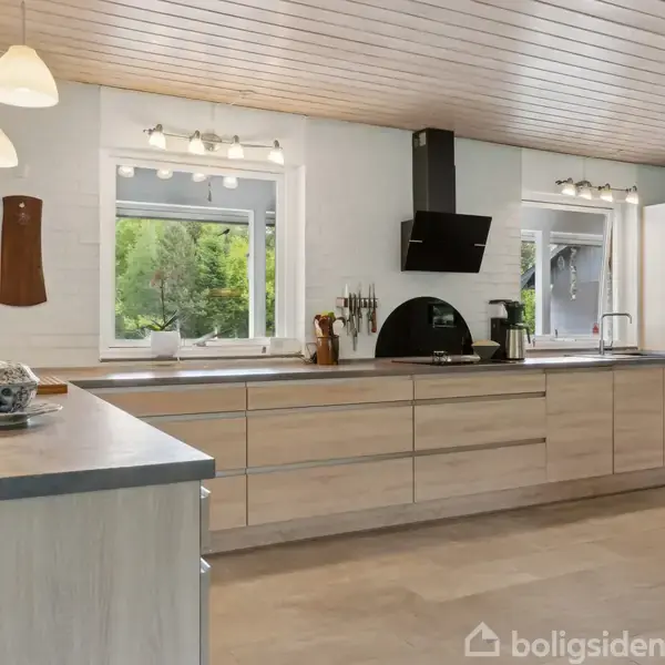 Et moderne køkken med træskabe og sort emhætte, oplyst af vinduer og lamper, set mod en grøn udendørs baggrund udenfor. Bordet er dækket med lys og dekorationer.