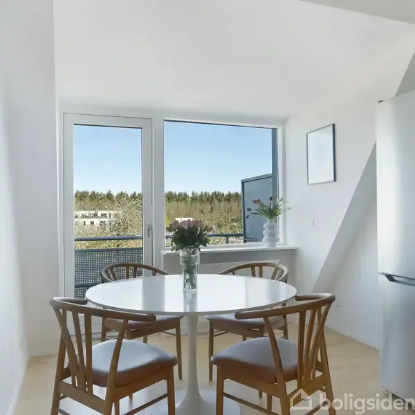 Et rundt spisebord er omringet af fire stole og har en vase med blomster, placeret i et lyst rum med store vinduer og udsigt til træer.