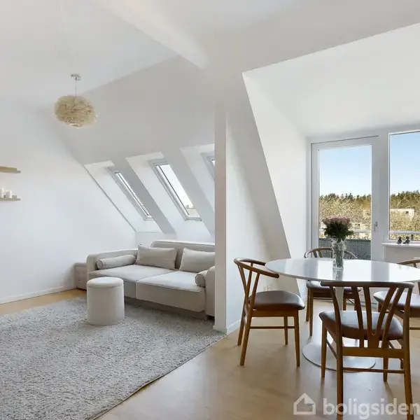 Stue med lyse møbler, herunder en sofa og et rundt spisebord med stole. Store vinduer i baggrunden giver udsigt til en skov. Hylder med dekorationer hænger på væggen.