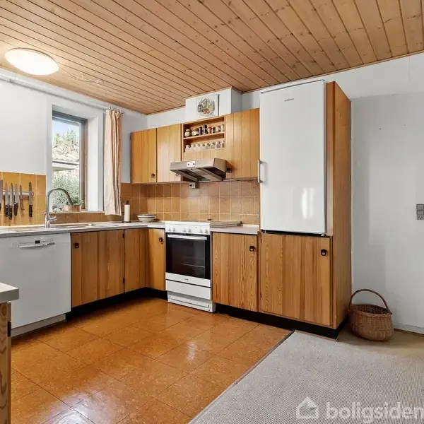 Køkken med træskabe indeholder en opvaskemaskine, komfur og køleskab. Skarpe knive hænger på væggen, lidt bordplads. Vindue giver udsyn til have og dagslys. Ved siden af ligger en åben dør til et andet værelse.