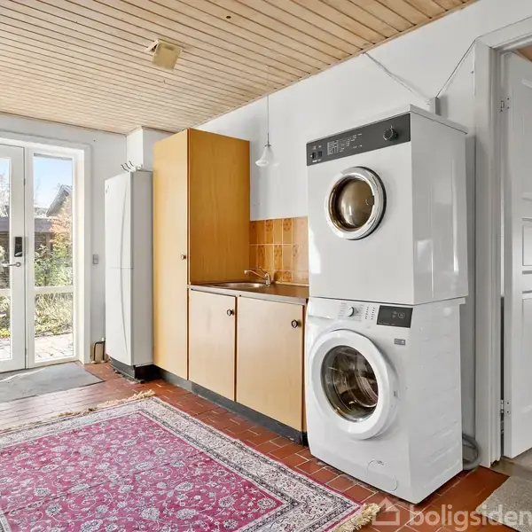 Vaskemaskine og tørretumbler står stablet i et bryggers med rødt flisegulv. Ved siden af er der en vask på et træbord. En stor dør åbner ud til en have.