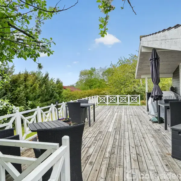 Terrasse lavet af træbrædder med sorte havemøbler under en parasol. Den ligger ved et hus med en overdækket del. Omgivet af grønne buske og træer under en klar himmel.
