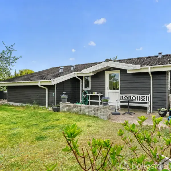 Sort træhus med lyse detaljer står stille i en grøn have med buske og græsplæne under en blå himmel. En veranda med hvid bænk er synlig ved siden af indgangen.