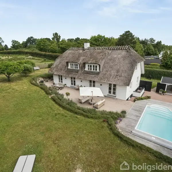 Stråtækt hus står stille i en frodig have med velplejet græsplæne og en rektangulær swimmingpool. Omkringliggende natur og terrasser skaber en fredelig atmosfære.