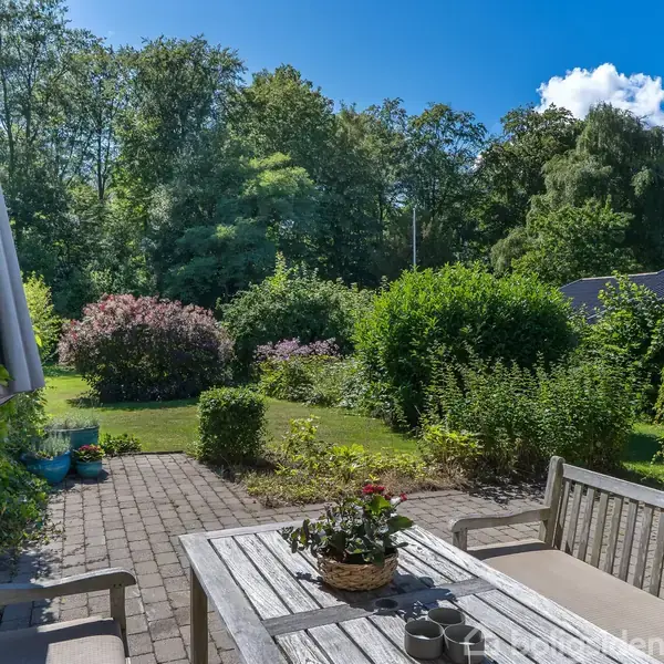 Træhavebord med stole står på flisebelagt terrasse. Bordet har en blomsterdekoration og tre kopper. Omgivet af frodig have med buske og træer under en blå himmel.