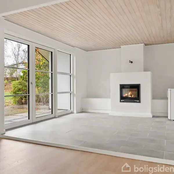 Pejs i funktion opvarmer rummet. Stuen har hvide vægge, lyse trægulve, og et gråt fliseområde omkring pejsen. Store vinduer til venstre åbner ud mod en have.