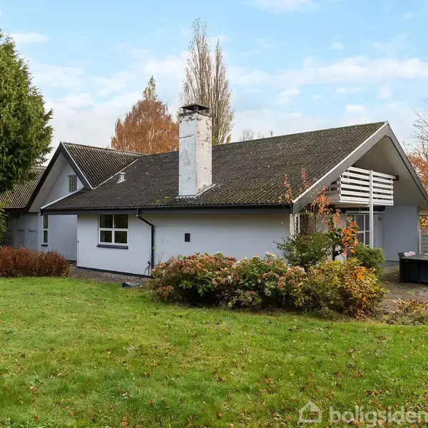 Et hvidt hus med sort tag står i en velplejet have. Huset har en skorsten, omkranset af grønne buske og træer. Baggrund med spredte, høje træer.