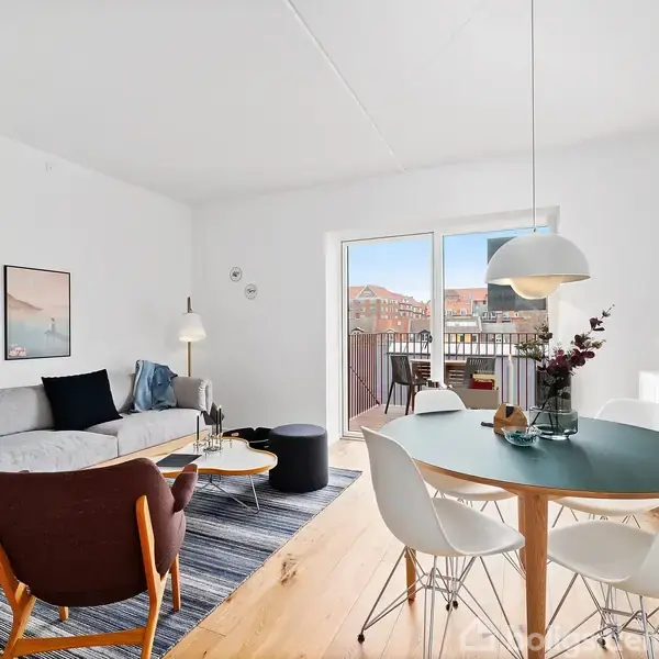 En stue indeholder en sofa foran et sofabord med lamper og kunst på væggen. Et rundt spisebord med stole står ved en stor altandør. Lys træparketgulv.