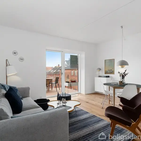 Stue med grå sofa, der står ved et lavt bord, vendt mod store vinduer med udsigt til en altan. Til højre står et rundt spisebord med stole under en lampe.