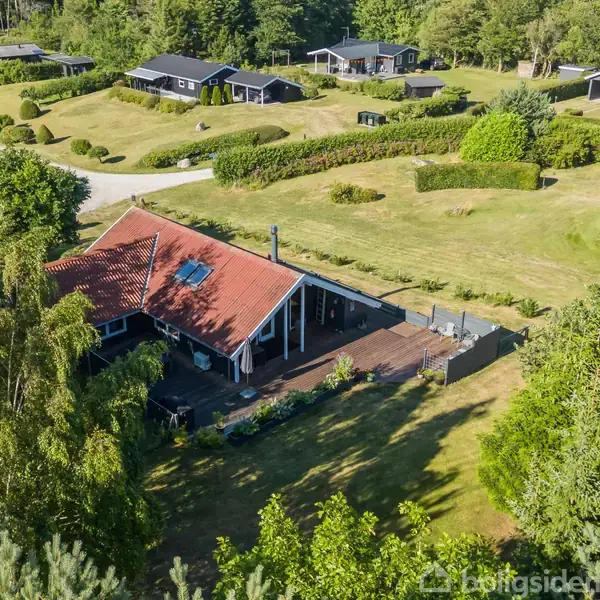 Rødt taghus med terrasse står blandt træer og buske. Omkringliggende haveområder og andre huse ses i baggrunden. Sommerhusområde præget af frodig natur.
