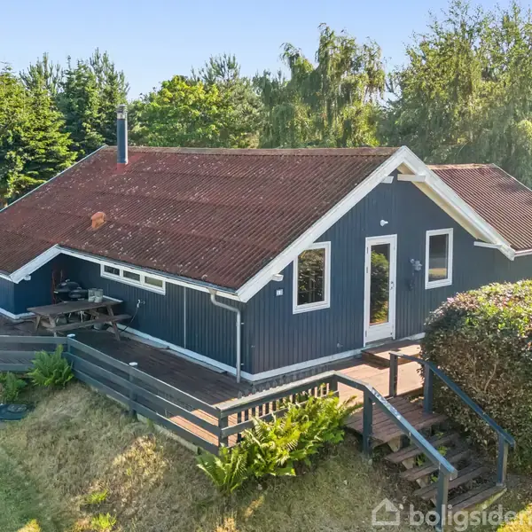 Et sommerhus med blå facade og rødt tag står omgivet af træer og græs. En terrasse med bord og stole ses til venstre.