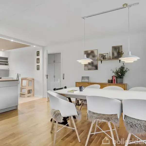 Spisebord med hvide stole står i et lyst spiseområde. Bordet er dækket med skindhynder. Køkkenet til venstre har en ø med lys træoverflade og vindue bagved.