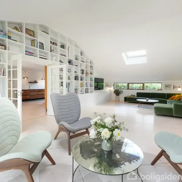En elegant stue med tre lysegrønne stole omkring et glasbord fyldt med blomster. Reoler fyldt med bøger og kunst dekorerer væggen, mens et grønt sofaarrangement står ved vinduerne.