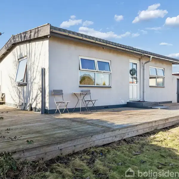Et enkelt hus med fladt tag står på en træterrasse. To stole er placeret ved siden af indgangsdøren. Omkring huset ses græs og buske under en blå, skyet himmel.