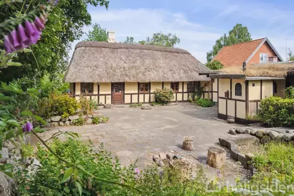 Bindingsværkshus med stråtag står i en gårdhave omgivet af grønne planter og blomster. Stenbelægning dækker gårdhaven, og en anden bygning med rødt tag ses i baggrunden.