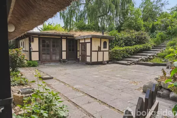 Halvbindingsværkshus med stråtag står stille i en brolagt gårdsplads. Haven omkring er grøn og frodig, med stenbelagte trin op mod et skrånende terræn, omgivet af buske og træer.