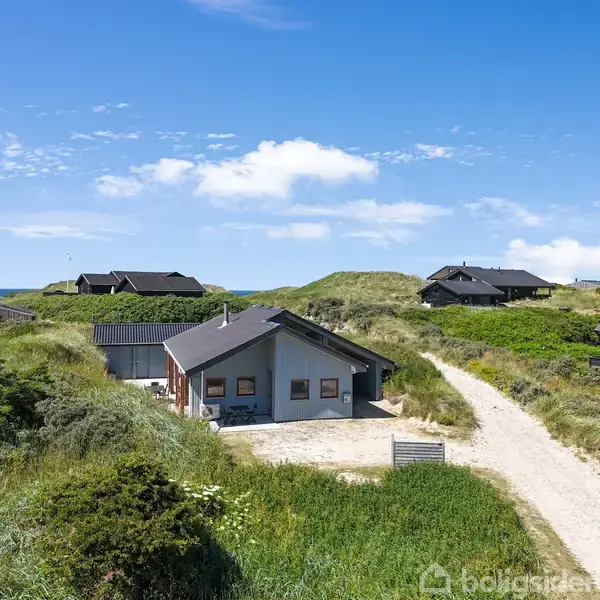 Et sommerhus står i klitterne, omgivet af græs og sandstier under en klar blå himmel. Flere huse ses i baggrunden blandt klitterne.