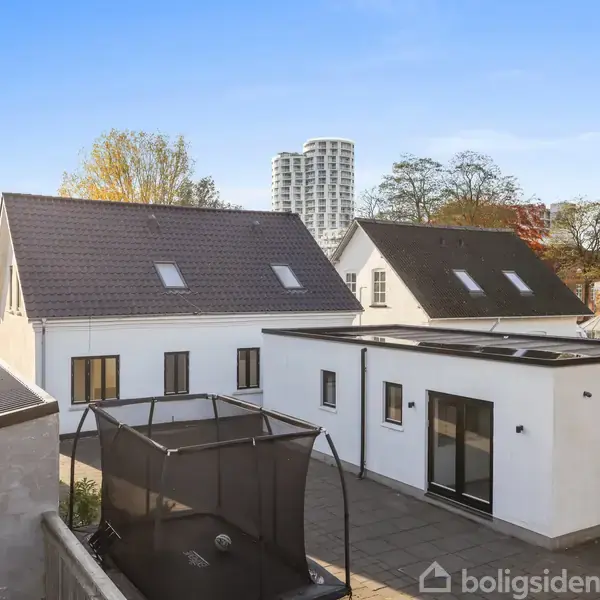 Hus med hvid facade står på en flisebelagt terrasse. En trampolin er placeret på terrassen. I baggrunden ses flere huse og højhuse mod en klar blå himmel.
