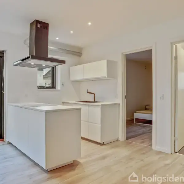 Køkken med hvide skabe og kogeø står i et lyst rum. En emhætte hænger over kogeøen. Døre fører til et soveværelse og badeværelse. Udsigt til gårdhave.