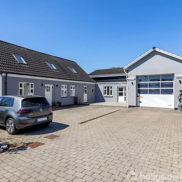 Gråt hus med skråtag står i en gårdsplads; bilen er parkeret i stenbelagt indkørsel. Garage med hvid port til højre.