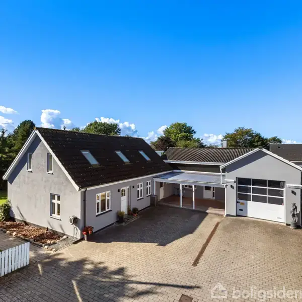 Et gråt hus med tilhørende garage står på en stor gårdsplads med brosten. Omkring huset er der en grøn have med træer, og himlen er klar og blå.