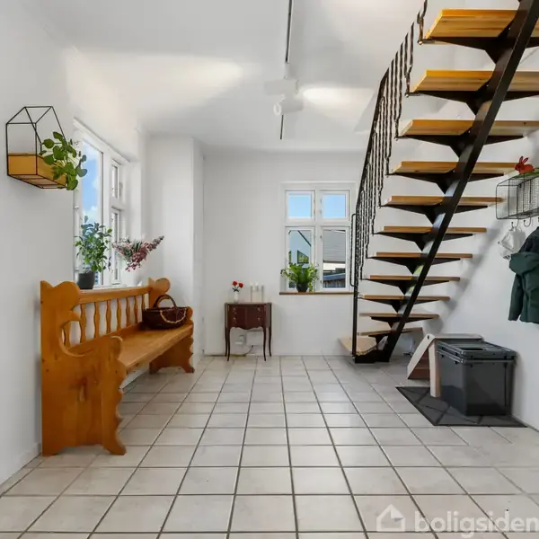 Træbænk står ved vindue i en lys entré med flisegulv; trappe fører op til næste etage. Væggen har frakkekroge med jakker. Indretningen er enkel og moderne.