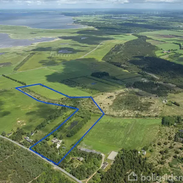 Stråtækt hus med røde mursten og hvide vinduer står på en gårdsplads med brosten. En træbænk og bord er placeret foran huset. Grønne træer omkranser området.