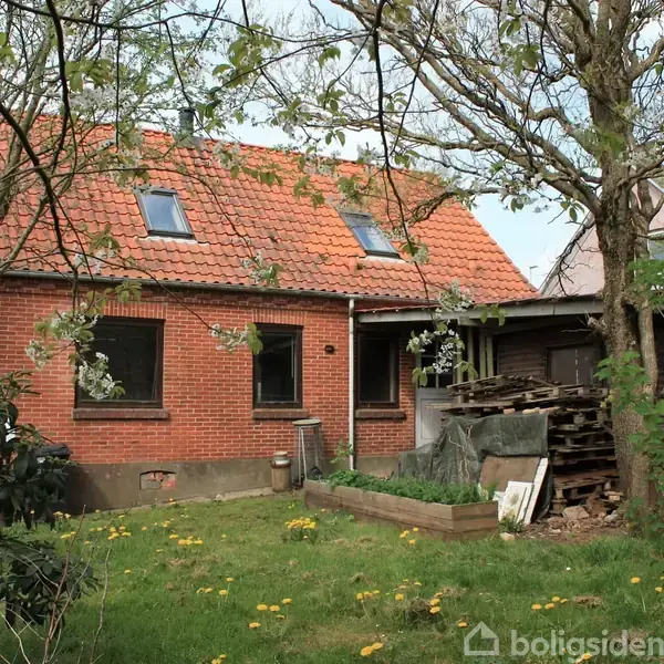 Et rødt murstenshus med skråt tegltag og tagvinduer. Omgivet af en grøn have med træer og blomster. Ved siden af huset ligger en bunke brænde og en lille skur.