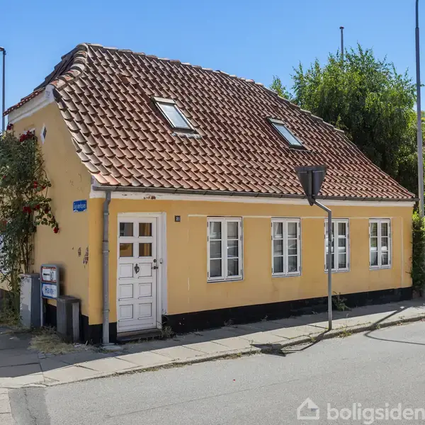 Gul murstensbygning med rødt tegltag og hvide vinduer, står ved vejkrydset. Et skilt med teksten "Lille Dam 2" ses ved siden af hoveddøren. Solrig dag.