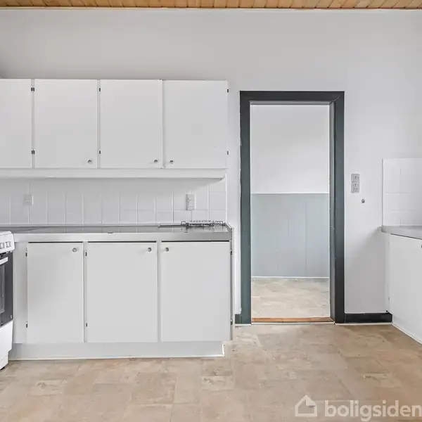 Et lyst køkken med hvide skabe og en ovn. Rummet har træloft og en døråbning fører til et tilstødende rum. Gulvet er lavet af lyse fliser.