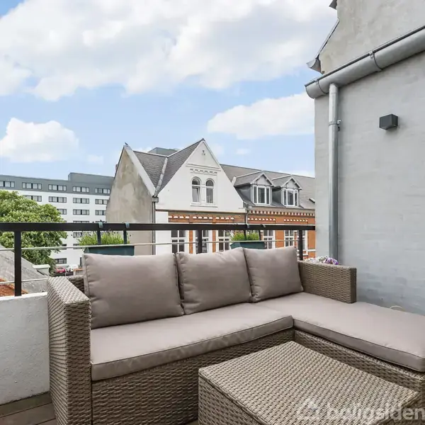 En stor træterrasse strækker sig langs en bygning med udsigt over byens tage. I baggrunden findes moderne bygninger og et blåt himmellandskab med skyer.