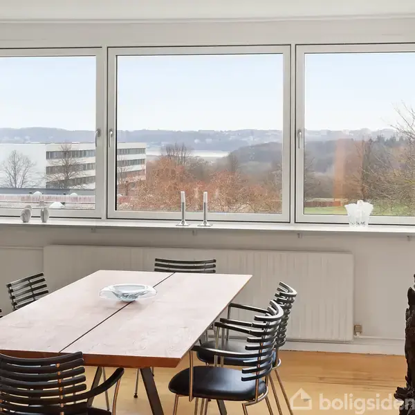 Et spisebord med sorte stole står foran store vinduer, der åbner op til en udsigt over et bakket landskab og en sø. Der er et kunstværk til venstre.