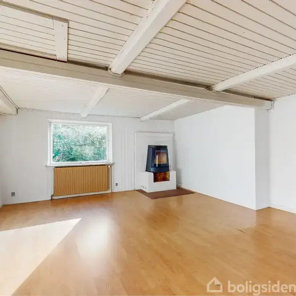 Stort, lyst rum med brændeovn placeret ved en hvid væg. I baggrunden er der et vindue med udsigt til træer og en radiator nedenunder. Loftet har synlige bjælker.