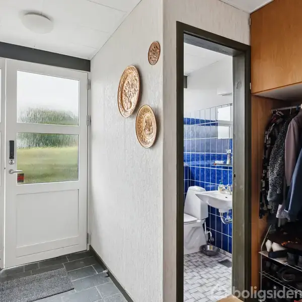 Døråbning med glaspartier fører til lyst rum med klinkegulv. Venstre side har kroge og jakker. Til højre ses et skab med tøj og sko samt en dør til et badeværelse med blå fliser.