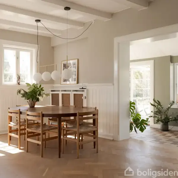 Rundt spisebord med stole står på trægulv. Bordet har en plante ovenpå. Store vinduer og lyse vægge skaber en lys atmosfære. Adgang til et rum med planter.