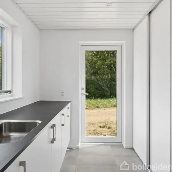 Et lyst køkken med en sort bordplade og vask. En glasdør fører til en udendørs have med træer. Bordet har hvide skabe og skuffegreb.