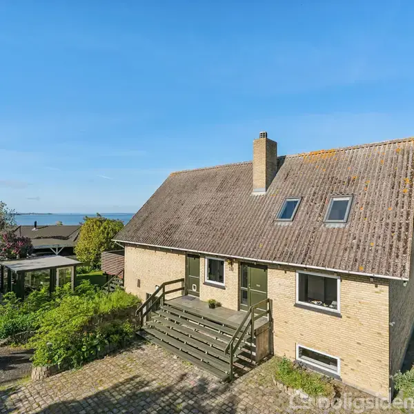 Et rummeligt, murstenshus med en stejl, mørk tagbelægning. Det står omgivet af frodig beplantning og en brolagt terrasse. I baggrunden ses et åbent marklandskab og havudsigt under en klar blå himmel.