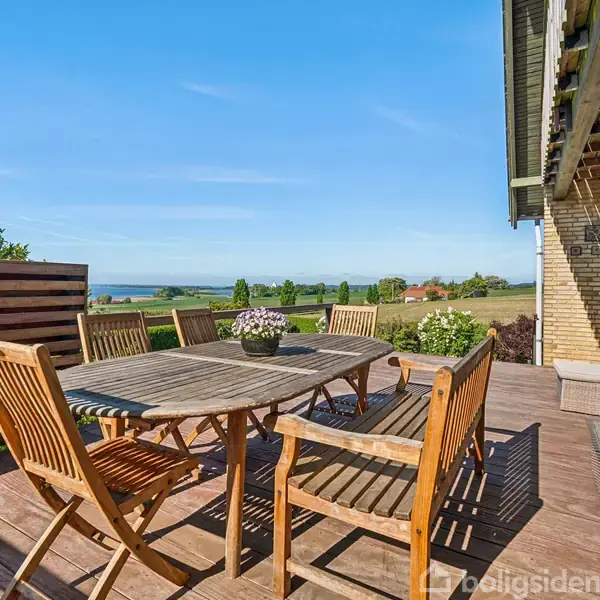 Træbord omgivet af stole står på en terrasse med udsigt over grønne marker og en fjern horisont. Vejret er solrigt med en klar blå himmel.