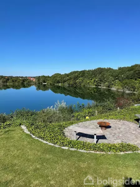 En rund bålplads med en bænk står stille ved en sø omgivet af frodige buske og træer under en klar blå himmel.