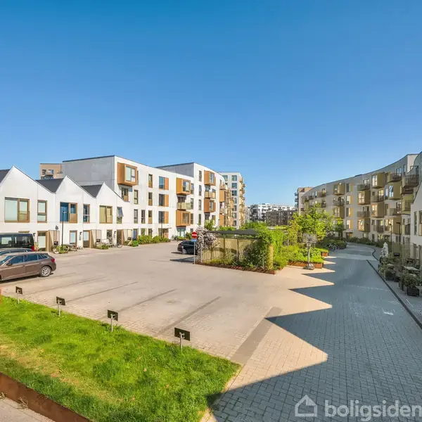 Beboelsesbygninger omkranser en asfalteret parkeringsplads med få biler, omgivet af grønne planter og moderne arkitektur, især karakteriseret ved hvide facader og mange vinduer. Klar blå himmel ses ovenfor.