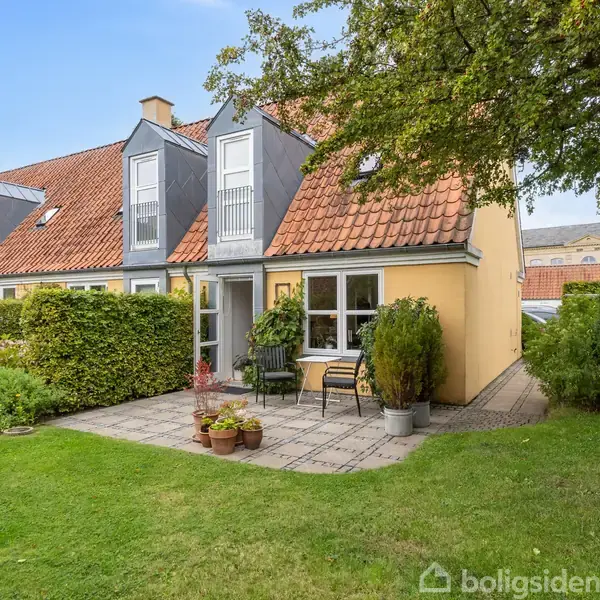 Et gult hus med rødt tegltag har en terrasse med havemøbler foran. Haven er grøn og omgivet af hække og træer, hvilket skaber en hyggelig atmosfære.